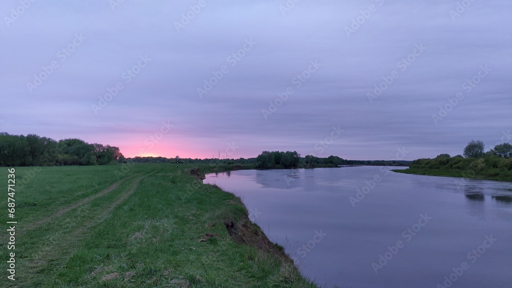 Spring evening, the sun has hidden behind the horizon. The river water reflects the cloudy pink-colored sky and trees. A forest grows on the opposite bank. On the water ripples. Cloudy
