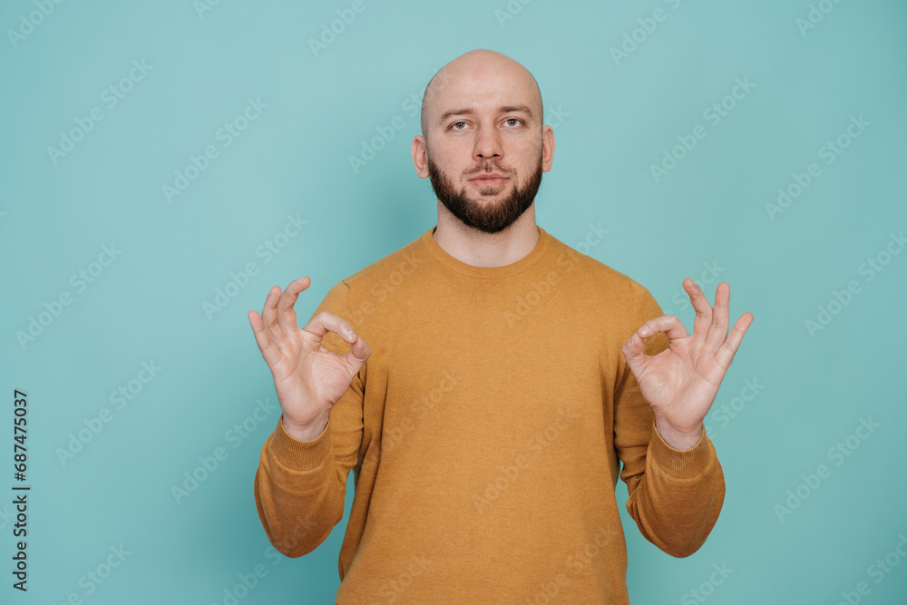 Fototapeta premium calm bald bearded man in casual holds hands in meditative gesture against turquoise background. focus concept. Mental balance concept. Keep calm.