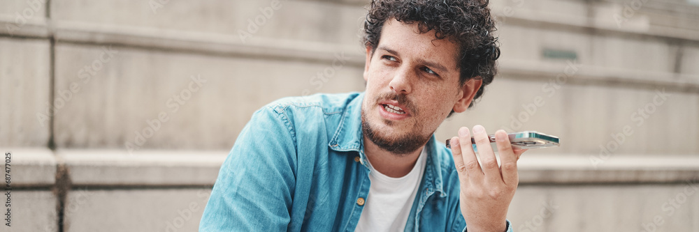 Fototapeta premium young bearded man in denim shirt sitting on high steps and using mobile phone. man sends voice message to smartphone