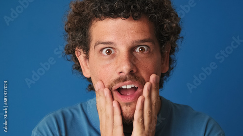 Close up, shocked guy with curly hair dressed in blue t-shirt looking in surprise at the camera with big eyes, raises his hands to his face, wow, isolated on blue background in the studio