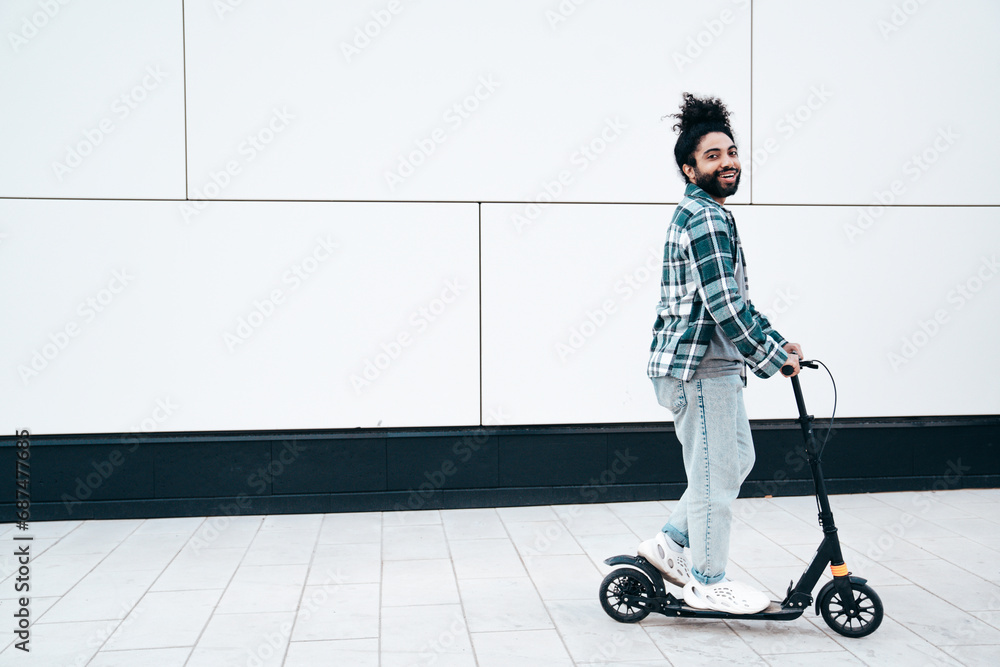 Trendy smiling bearded man in casual clothes riding electric scooter in urban background. Handsome model posing in the street near wall. Hipster guy with curly hairstyle. Cheerful and happy Trendy smiling bearded man in casual clothes riding electric scooter in urban background. Handsome model posing in the street near wall. Hipster guy with curly hairstyle. Cheerful and happy