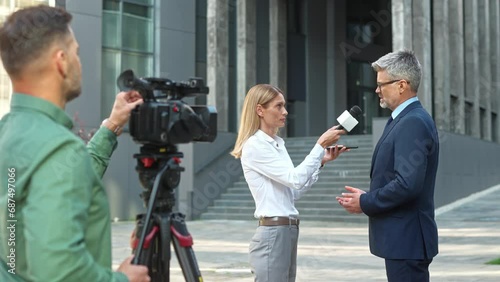 Busy cameraman recording interview of beautiful female journalist with famous politician in the middle of street. Pretty woman correspondent speaking with microphone to successful official.