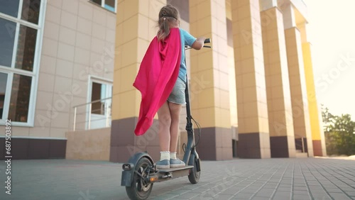 Girl superhero on electric scooter. happy family kid concept. a child on electric scooter rides on a large square in the city center dream under a blue sky with clouds and rays of sun. superhero girl