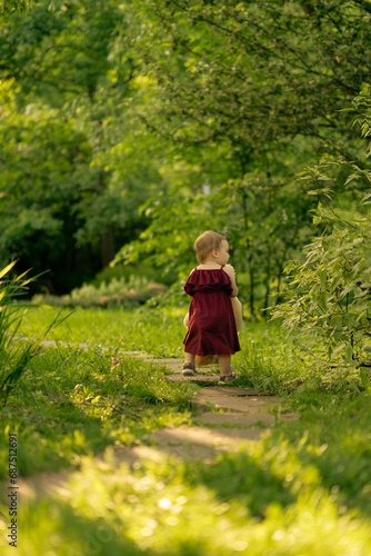 A small child in a maroon dress takes confident steps forward, her trusted teddy bear by her side, as they embark on an adventure down a garden path. © yavdat