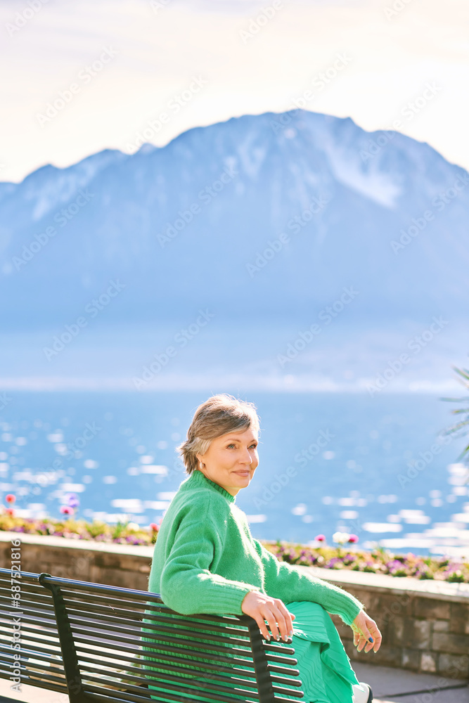 Fototapeta premium Portrait of middle age woman enjoying nice day outside, relaxing on bench in front of mountain lake