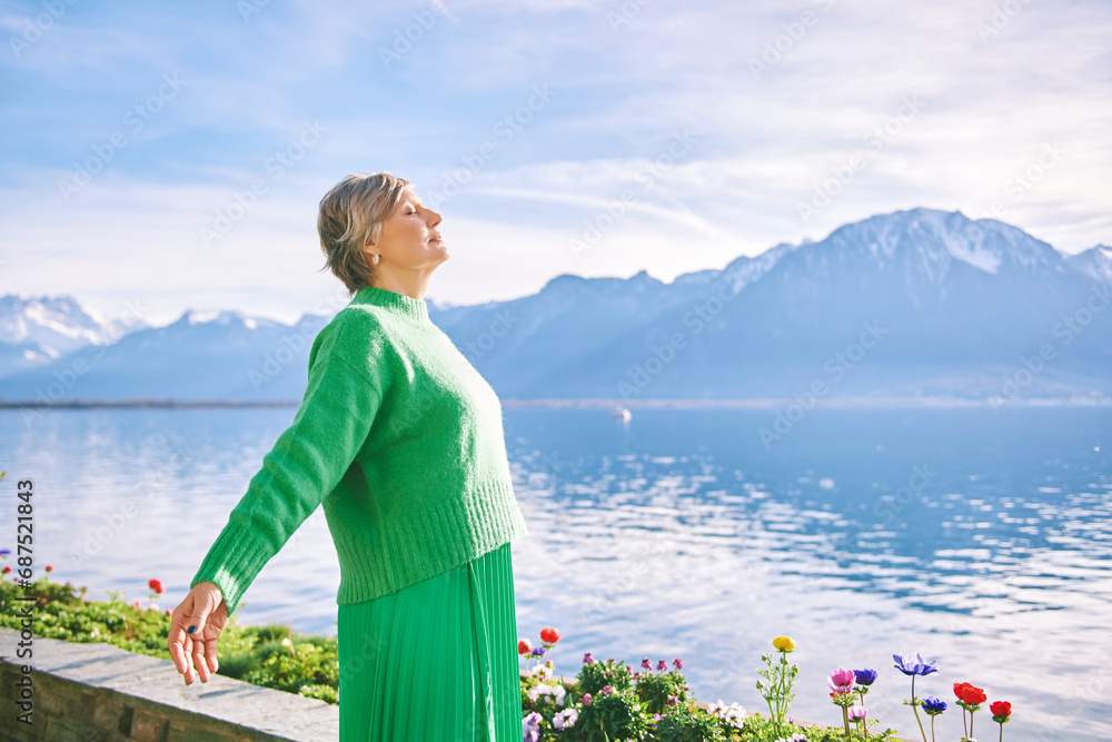 Fototapeta premium Outdoor portrait of happy middle age 55 - 60 year old woman with arms open wide, posing on blue sky and mountain background