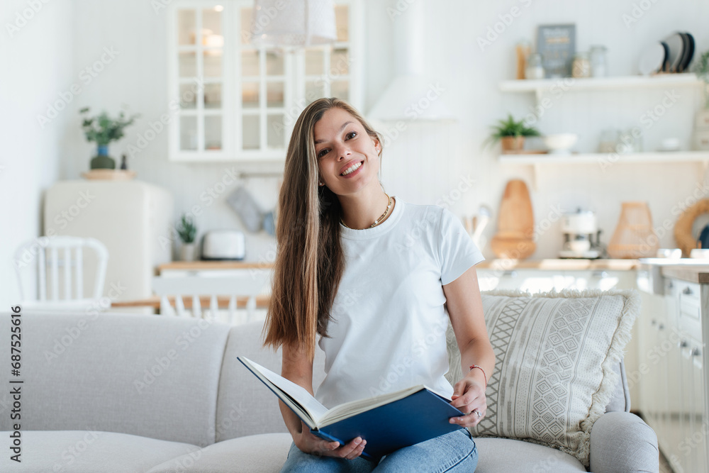 cheerful young girl with long hair in jeans and a white T-shirt is sitting at home on the couch reading a book and smiling on a sunny summer day. Rest at home, weekends cheerful young girl with long hair in jeans and a white T-shirt is sitting at home on the couch reading a book and smiling on a sunny summer day. Rest at home, weekends