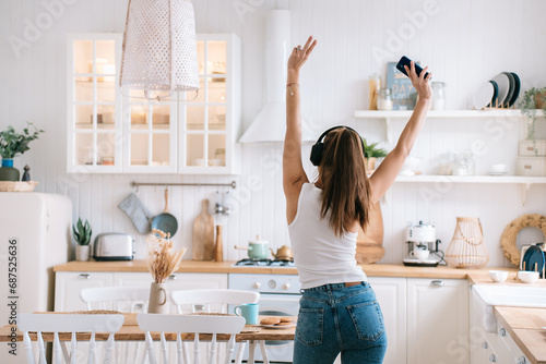 rear view of a positive girl in jeans dancing in the kitchen at home using headphones and a phone that raised her hands up shows a victory sign a fun weekend at home photo