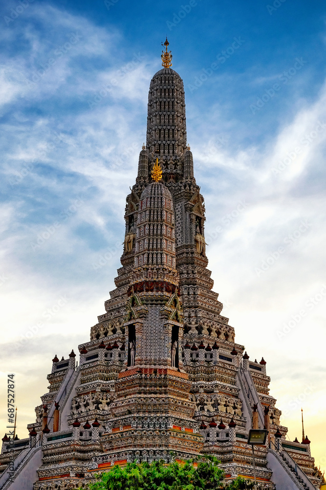 The Wat Arun temple in Bangkok, Thailand. The temple is a tall, ornate structure with a pointed spire. The temple is covered in intricate decorations and statues. The sky is blue with wispy clouds.