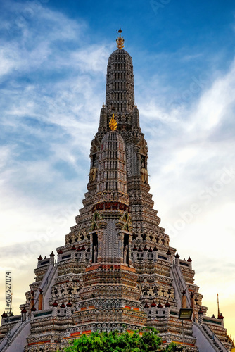 The Wat Arun temple in Bangkok  Thailand. The temple is a tall  ornate structure with a pointed spire. The temple is covered in intricate decorations and statues. The sky is blue with wispy clouds.