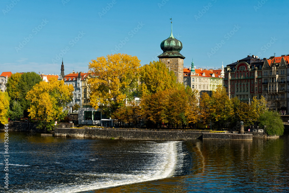A scenic view of a river with a waterfall and colorful autumn trees in the background. Ancient tower on the river bank in the fall. Autumn Prague. Landscape.