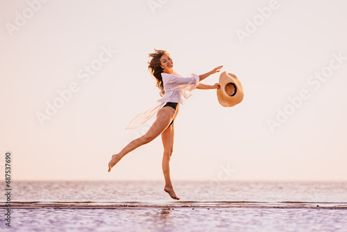 Tablou pe pânză Jumping high young gorgeous woman in black swimsuit with fluttering hair, white shirt rising hat up spending time on the beach smiles wide