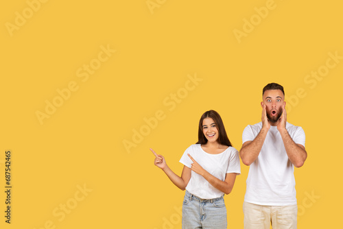 Glad shocked young european man with open mouth, woman in white t-shirts point finger at copy space