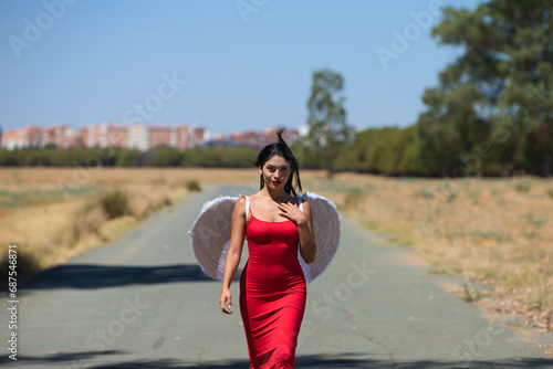 Beautiful young Latin woman in red silk dress and white wings walks along a tarmac road in the countryside outside the big city. The woman makes different poses and expressions.