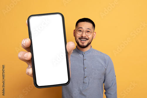 Asian man holding large smartphone with blank screen, yellow backdrop