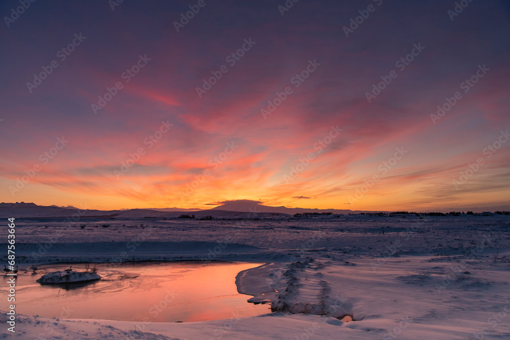 Panoramic view over a snowy landscape with river in Iceland during sunrise with the clouds coloring bright orange and yellow behind the mountains in the background and reflection in the water