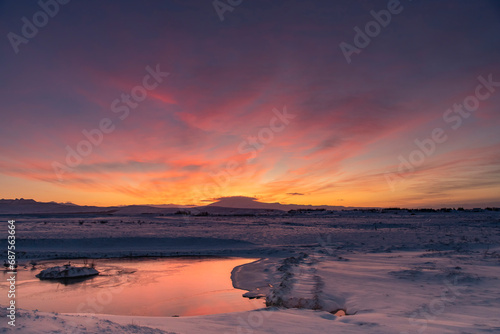 Panoramic view over a snowy landscape with river in Iceland during sunrise with the clouds coloring bright orange and yellow behind the mountains in the background and reflection in the water