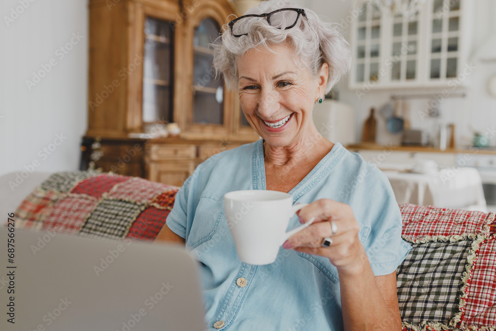 Side view of cute charming gray-haired grandma drinking coffee and watching favorite show on laptop or video chatting with grandchildren sitting on couch, relaxing and feeling carefree Side view of cute charming gray-haired grandma drinking coffee and watching favorite show on laptop or video chatting with grandchildren sitting on couch, relaxing and feeling carefree