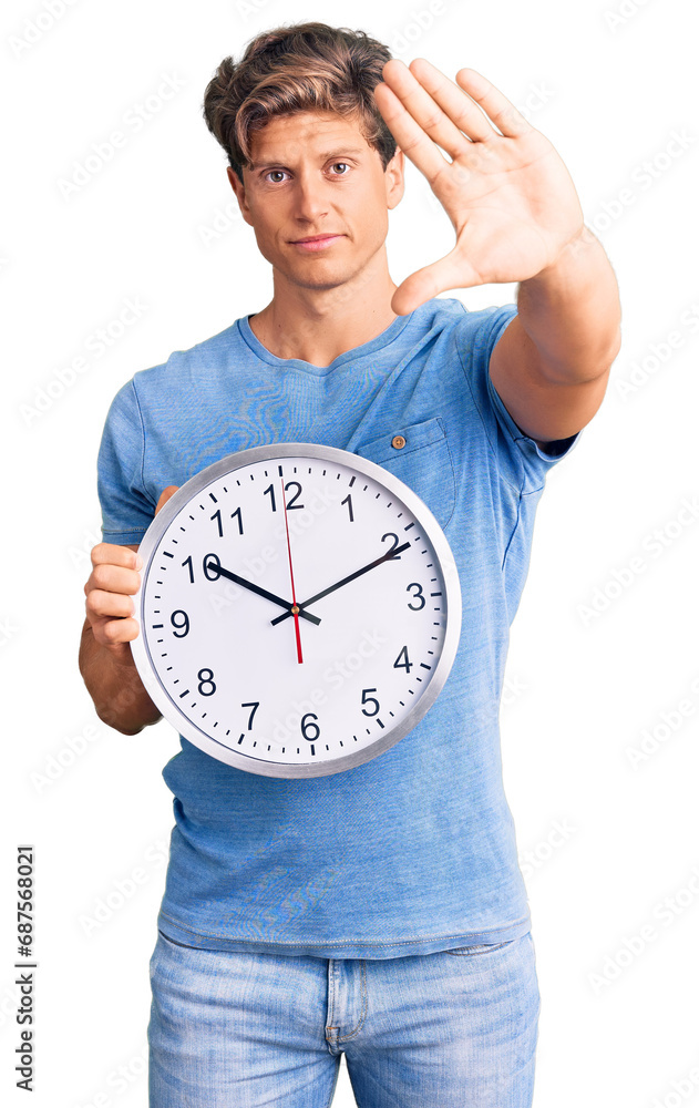 Young handsome man holding big clock with open hand doing stop sign with serious and confident expression, defense gesture Young handsome man holding big clock with open hand doing stop sign with serious and confident expression, defense gesture