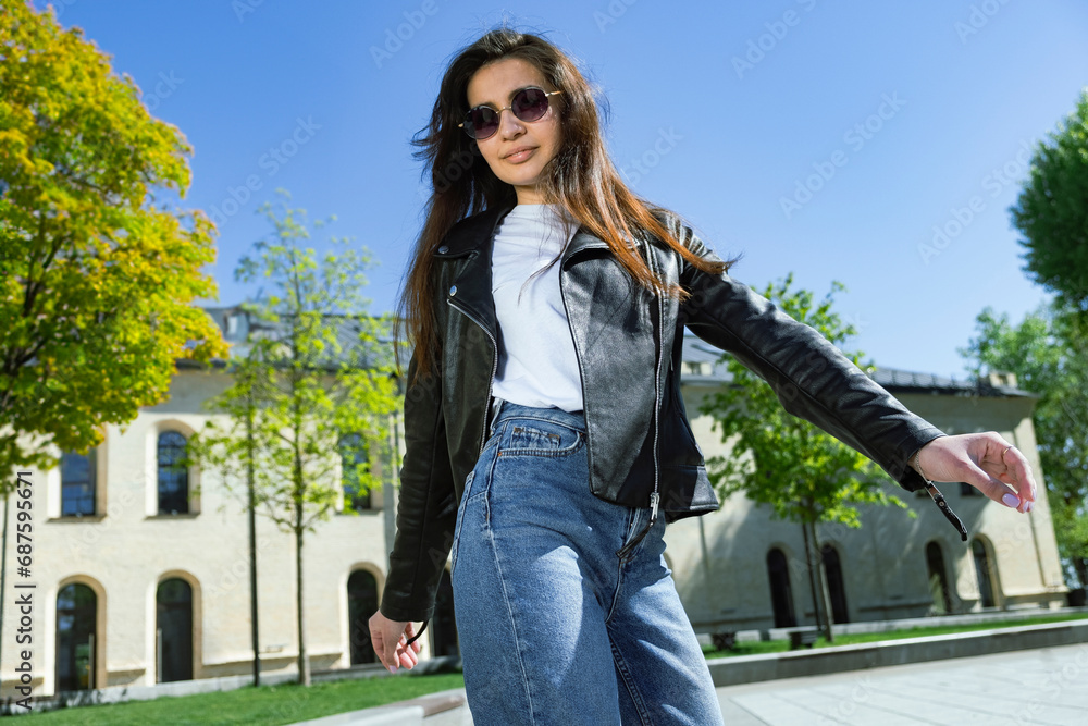 Fototapeta premium a brunette girl walks on the street in a leather jacket and sunglasses