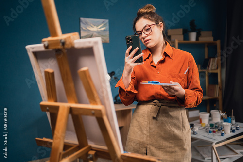 Happy young woman photographing her painting in art studio uses smartphone