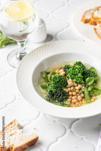 Vegetarian minestrone soup with green vegetables and beans served in white plate and bread on white tile background