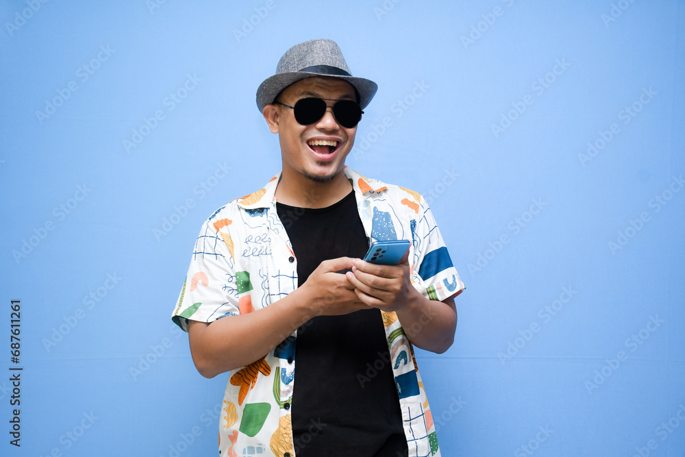 Happy asian man wearing beach shirt holding a phone while pointing a finger standing over blue background. Vacation concept