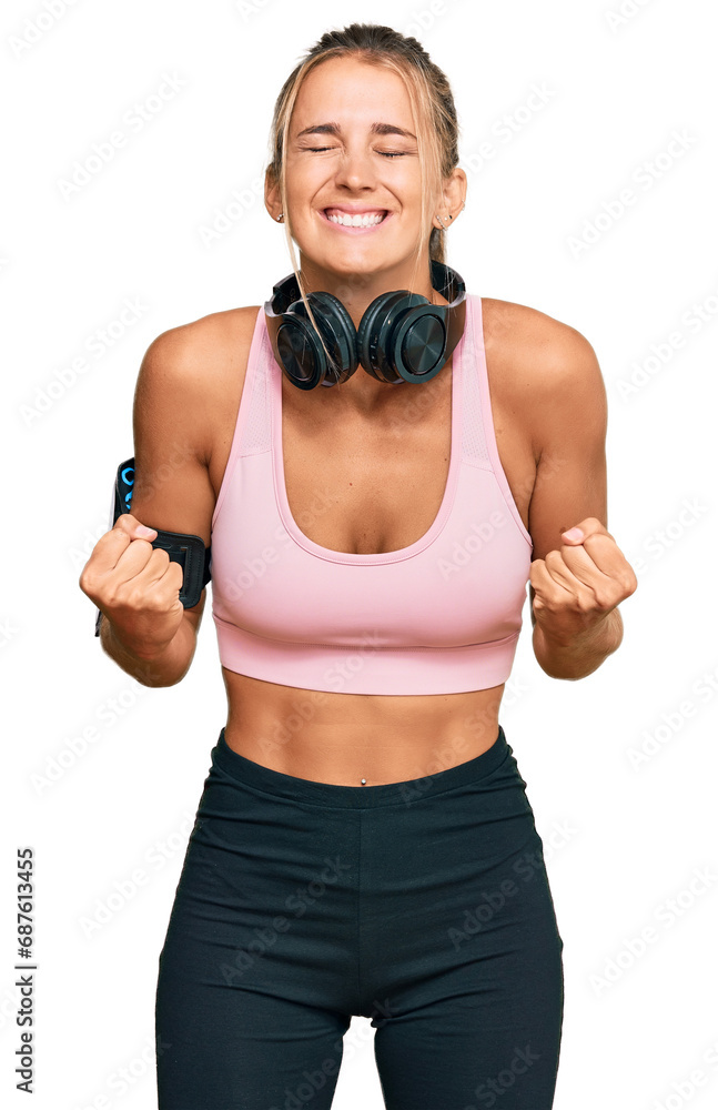 Young blonde woman wearing gym clothes and using headphones very happy and excited doing winner gesture with arms raised, smiling and screaming for success. celebration concept.