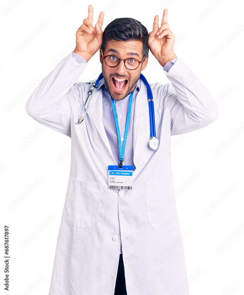 Young hispanic man wearing doctor uniform and stethoscope posing funny and crazy with fingers on head as bunny ears, smiling cheerful