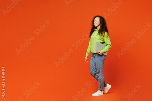 Full body side view confident young IT woman of African American ethnicity wears green hoody casual clothes hold closed laptop pc computer isolated on plain red orange background. Lifestyle concept.