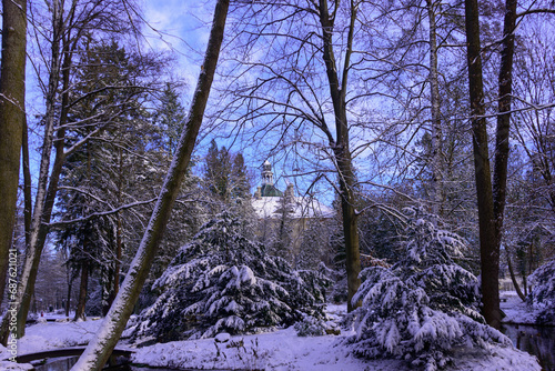 Pałac w parku zimą wśród drzew i krzewów są one pokryte warstwą śniegu, który pokrywa również ziemię. Niebo jest błękitne, lekko zachmurzone.