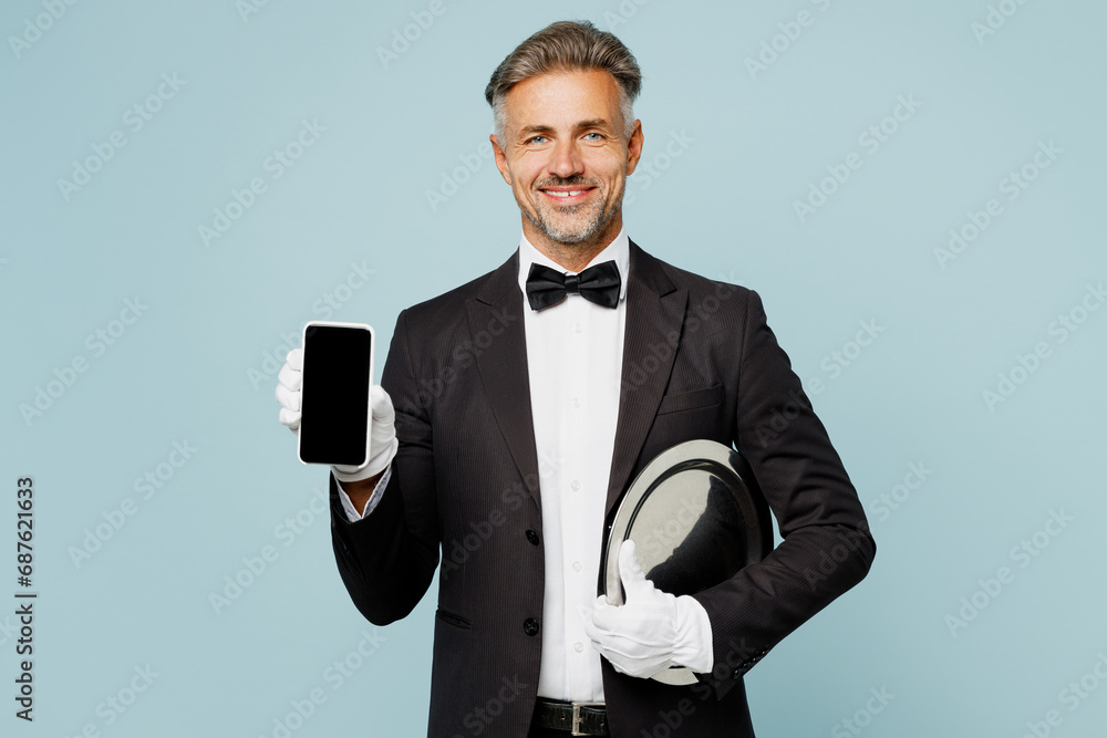 Adult barista male waiter butler man wear shirt black suit bow tie elegant uniform hold use blank screen mobile cell phone work at cafe isolated on plain blue background. Restaurant employee concept. - obrazy, fototapety, plakaty 
