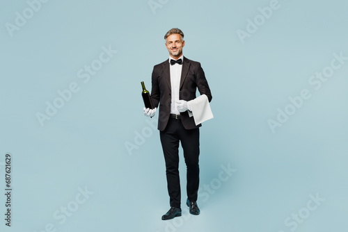 Full body adult sommelier barista male waiter butler man wears shirt black suit bow tie uniform hold in hand bottle of wine work at cafe isolated on plain blue background. Restaurant employee concept. photo
