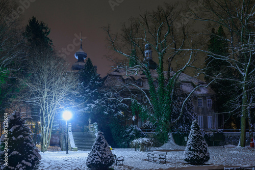 Pałac w parku zimą wśród drzew i krzewów są one pokryte warstwą śniegu, który pokrywa również ziemię. Jest noc, pałac jest skryty w mroku oświetlony nikłym światłem pobliskich latarni.