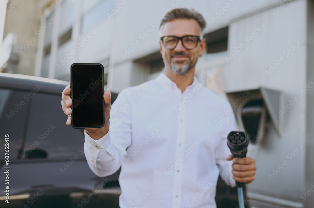 Adult man wears shirt use blank screen mobile cell phone hold in hand electric car charger connect to electric car auto at charging station outdoor. Green energy solution for automobile fuel concept. Adult man wears shirt use blank screen mobile cell phone hold in hand electric car charger connect to electric car auto at charging station outdoor. Green energy solution for automobile fuel concept.
