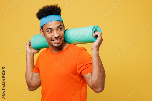 Young fitness trainer instructor sporty man sportsman wears orange t-shirt hold yoga mat look aside on area spend time in home gym isolated on plain yellow background. Workout sport fit abs concept.