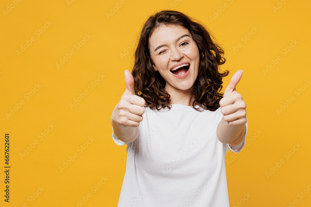 Young smiling happy cheerful Caucasian fun woman she wear white blank t-shirt casual clothes showing thumb up like gesture wink blink eye isolated on plain yellow orange background. Lifestyle concept. Young smiling happy cheerful Caucasian fun woman she wear white blank t-shirt casual clothes showing thumb up like gesture wink blink eye isolated on plain yellow orange background. Lifestyle concept.