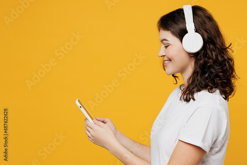 Side view young fun woman she wear white blank t-shirt casual clothes listen to music in headphones use mobile cell phone isolated on plain yellow orange background studio portrait. Lifestyle concept. photo