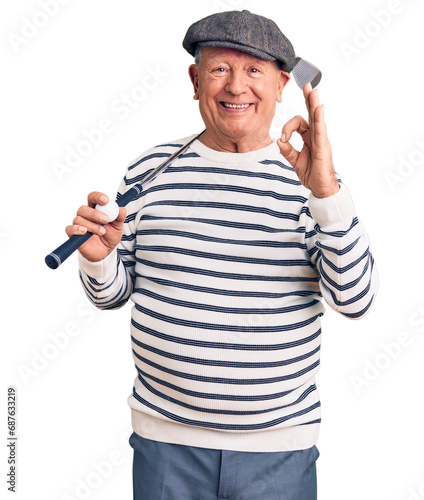 Senior handsome grey-haired man holding golf club and ball doing ok sign with fingers, smiling friendly gesturing excellent symbol