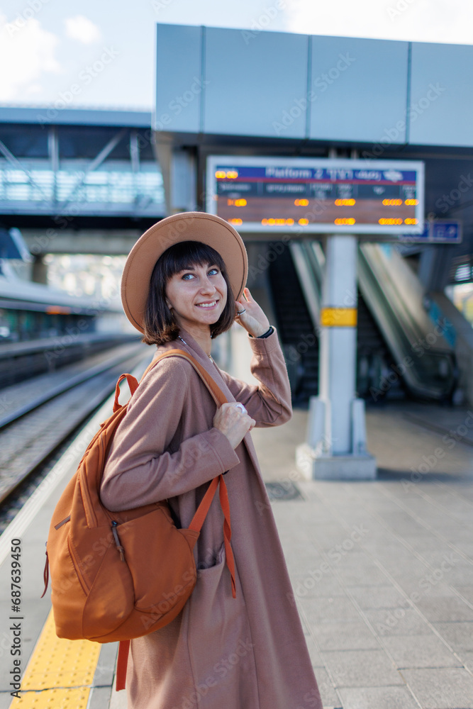 traveling by train. A young woman in a coat and hat stands at the station and looks at the train schedule.