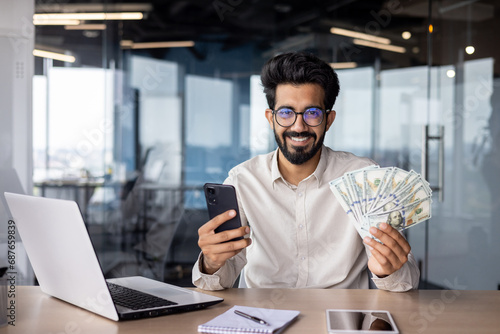 Portrait of a young Indian man sitting in the office at the table, holding cash money and a phone in his hands, smiling and looking at the camera photo