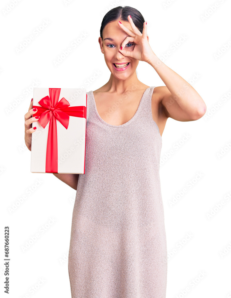 Beautiful brunette young woman holding gift smiling happy doing ok sign with hand on eye looking through fingers Beautiful brunette young woman holding gift smiling happy doing ok sign with hand on eye looking through fingers