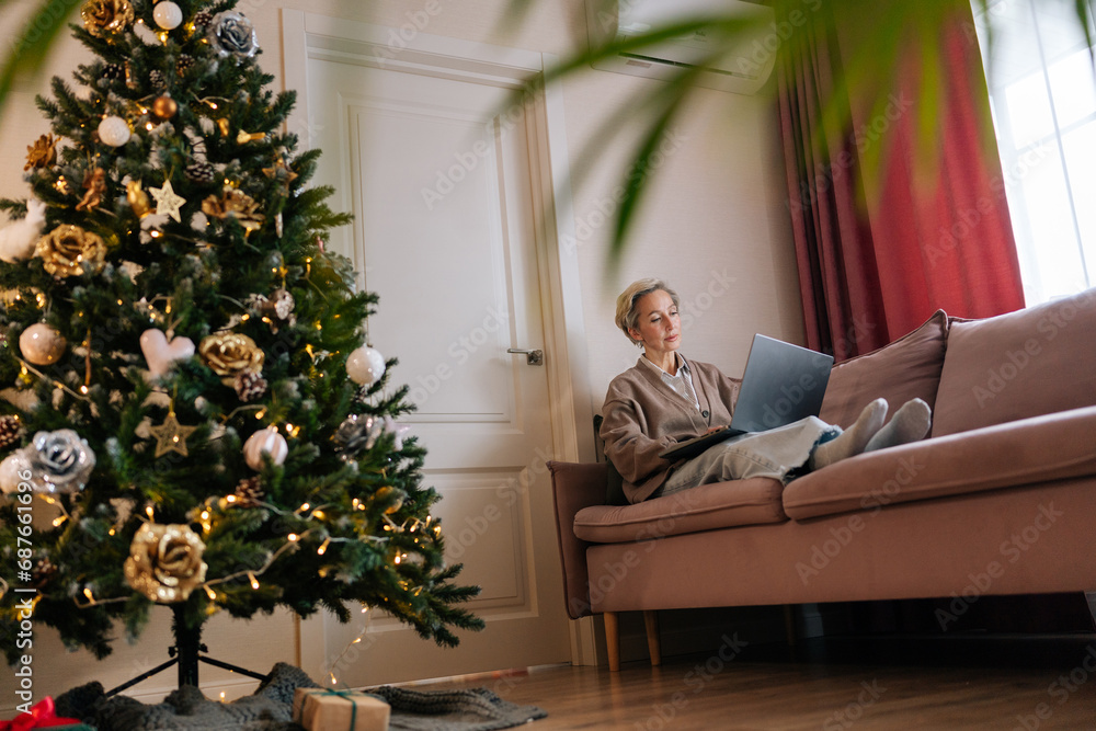 Remote view of pretty elegant middle-aged woman remote working or shopping online buying new year presents sitting on couch with laptop at Christmas time, with decorated Christmas tree on foreground. Remote view of pretty elegant middle-aged woman remote working or shopping online buying new year presents sitting on couch with laptop at Christmas time, with decorated Christmas tree on foreground.