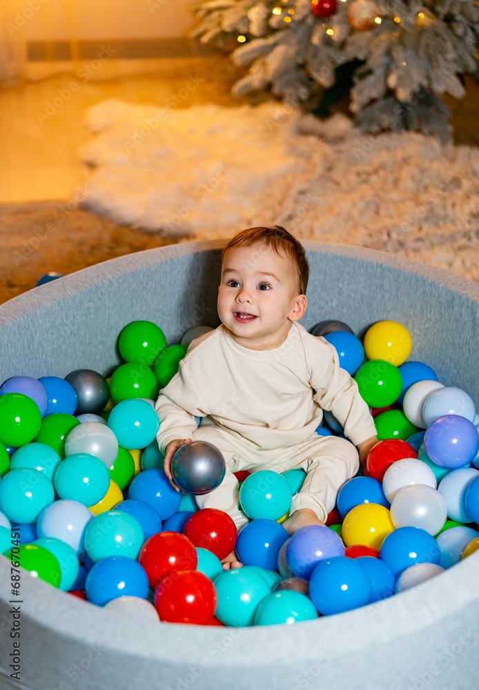 Obraz premium A Joyful Baby Surrounded by Colorful Balls, Gazing at a Festive Christmas Tree. A baby sitting in a ball pit with a christmas tree in the background