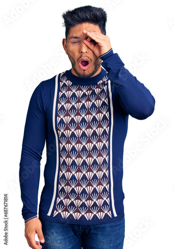 Handsome latin american young man wearing casual winter sweater doing ok gesture shocked with surprised face, eye looking through fingers. unbelieving expression.