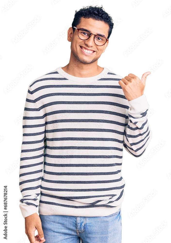 Hispanic handsome young man wearing casual clothes and glasses smiling with happy face looking and pointing to the side with thumb up. Hispanic handsome young man wearing casual clothes and glasses smiling with happy face looking and pointing to the side with thumb up.