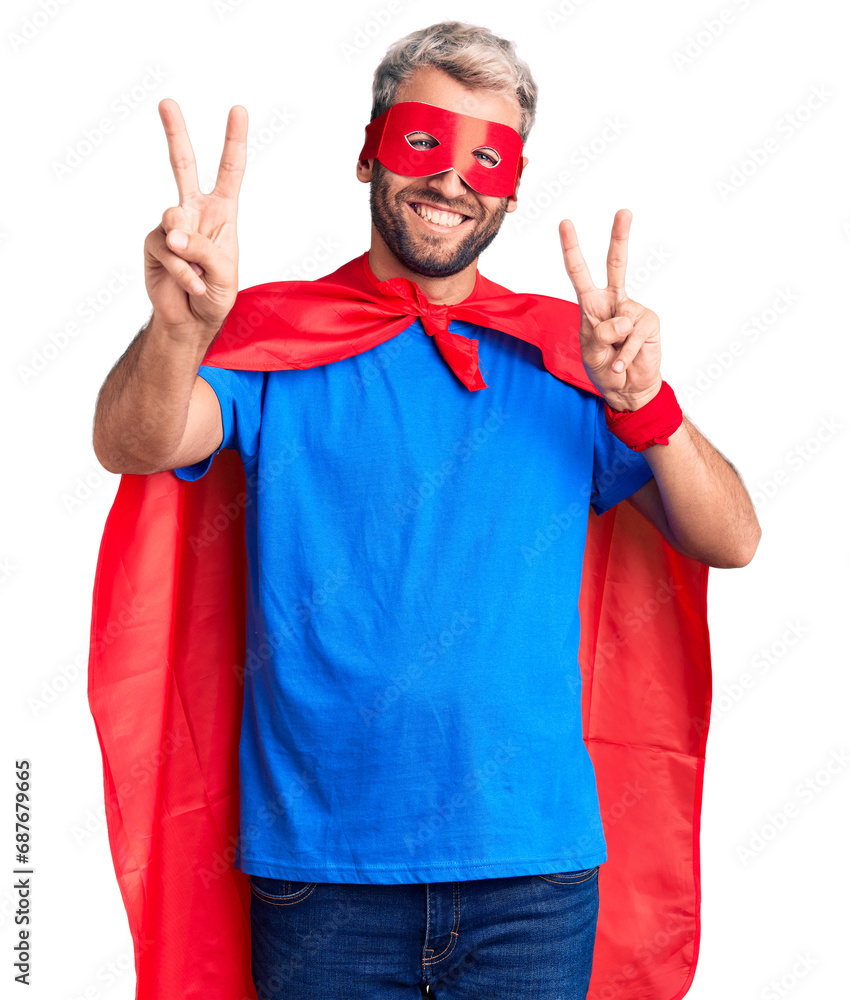 Young blond man wearing super hero custome smiling looking to the camera showing fingers doing victory sign. number two. Young blond man wearing super hero custome smiling looking to the camera showing fingers doing victory sign. number two.