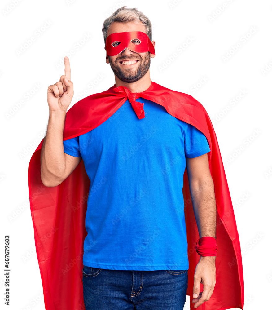 Naklejka premium Young blond man wearing super hero custome showing and pointing up with finger number one while smiling confident and happy.