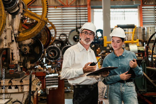 Factory engineer manager with assistant using laptop to conduct inspection of steel industrial machine, exemplifying leadership as machinery engineering inspection supervisor in metalwork manufacture.