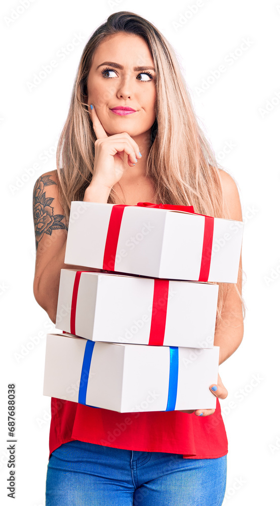 Young beautiful blonde woman holding birthday gifts serious face thinking about question with hand on chin, thoughtful about confusing idea Young beautiful blonde woman holding birthday gifts serious face thinking about question with hand on chin, thoughtful about confusing idea